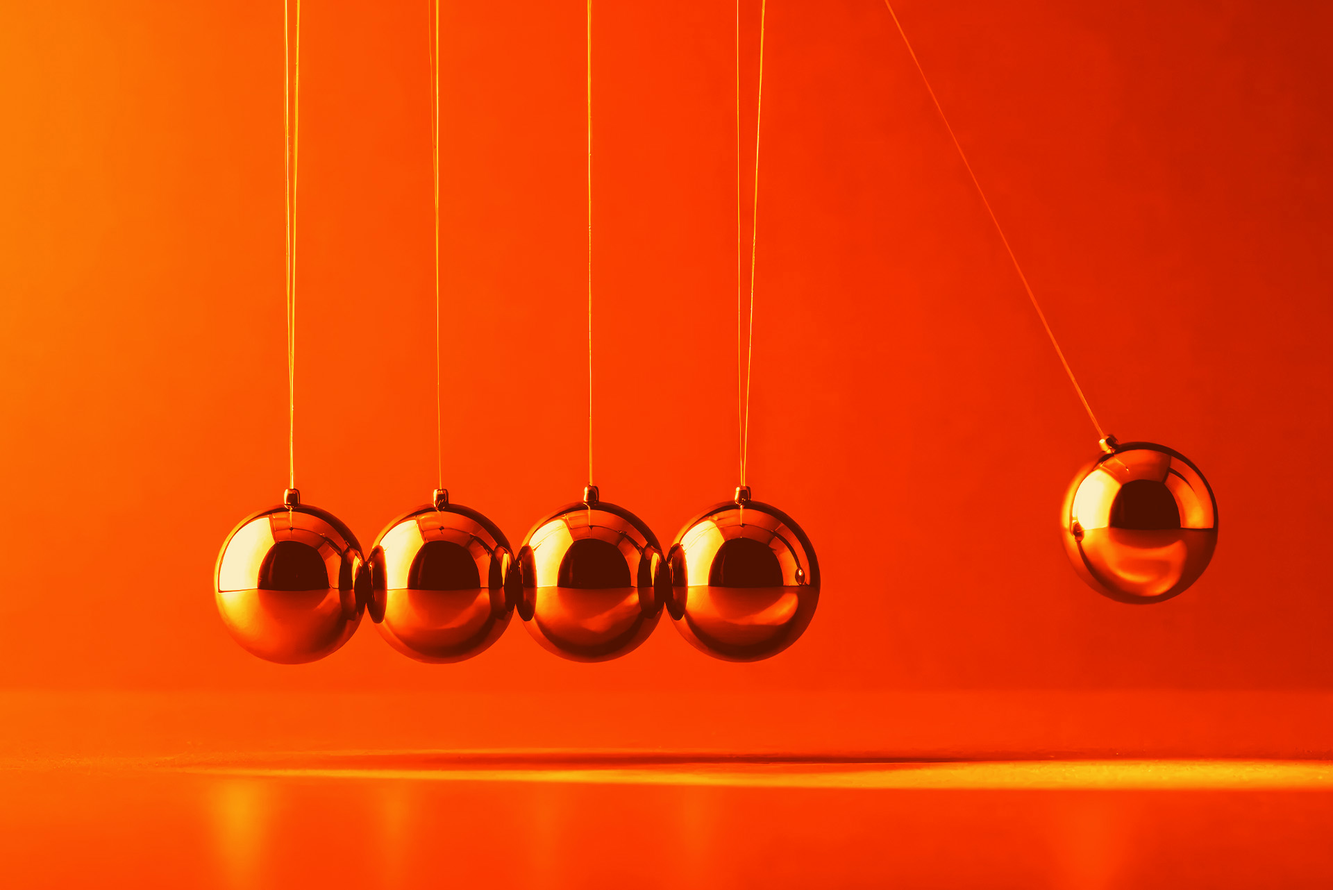 Five metallic pendulum balls suspended against a vibrant orange background, with one ball swinging away from the others.
